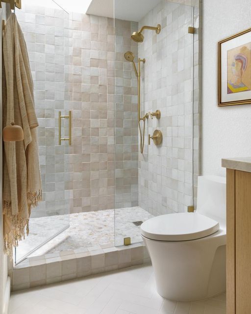 a catchy and light-filled neutral bathroom clad with neutral Zellige tiles, a skylight over the shower, white appliances and gold fixtures