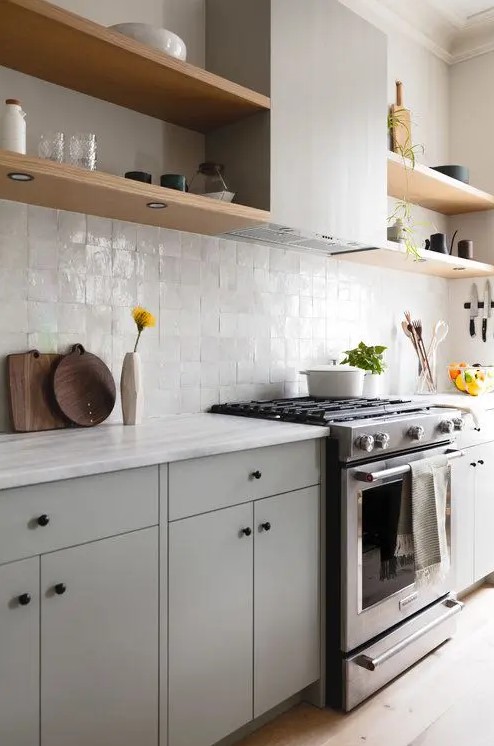 A chic dove grey kitchen with a white zellige tile backsplash, light stained shelves and white stone countertops is amazing