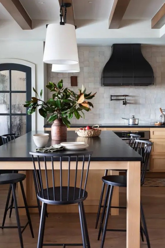 A chic vintage inspired kitchen with stained shaker cabinets and a large table kitchen island, black statement countertops, a black hood and black chairs