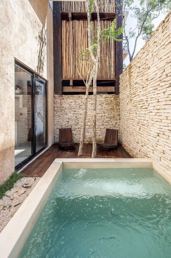A minimal outdoor space with stone walls, a dark stained furniture, a plunge pool, some trees and wooden chairs