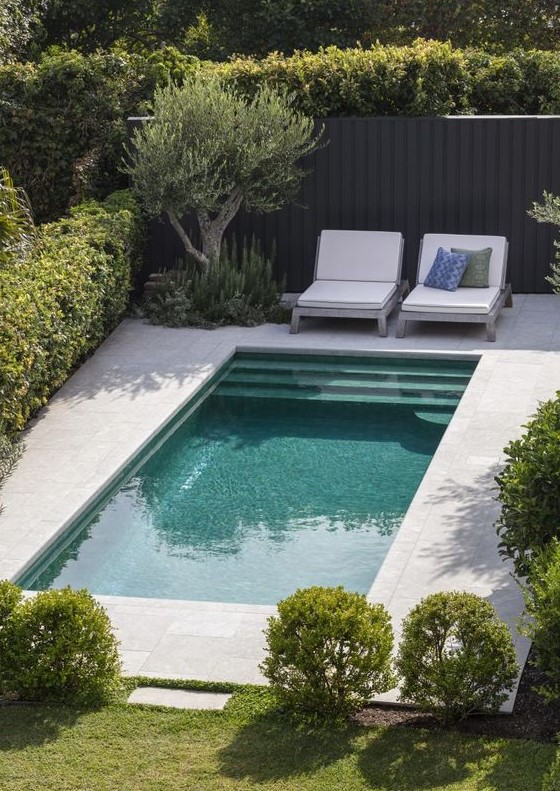 a minimalist backyard with a green lawn and bushes, with a pool and a stone deck and a couple of chairs is a lovely space