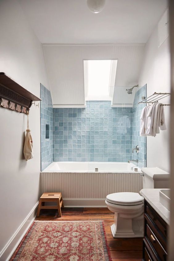 a modern bathroom with blue squre tiles around the tub and a skylight over the tub, a dark-stained vanity, a bold rug and some neutral fixtures