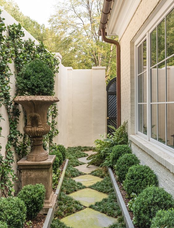 a beautiful vintage side yard with a pavement path and greenery, shrubs and a vintage urn with greenery on a stand