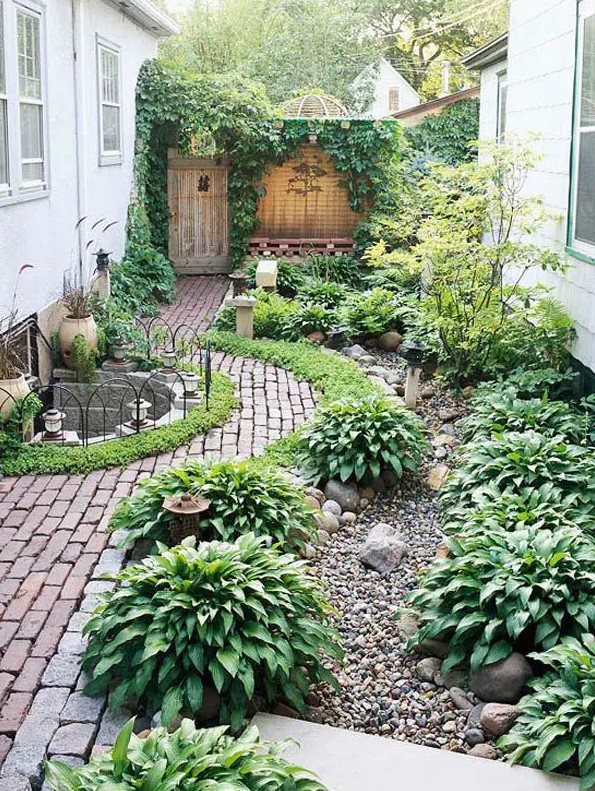 a lush green side yard with a lot of plants, brick paths, gravel and potted plants, too, is a very cozy and lovely space