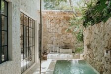 17 a tropical outdoor space with a stone fence, a narrow pool with a waterfall, a seat with pillows and some trees over the space