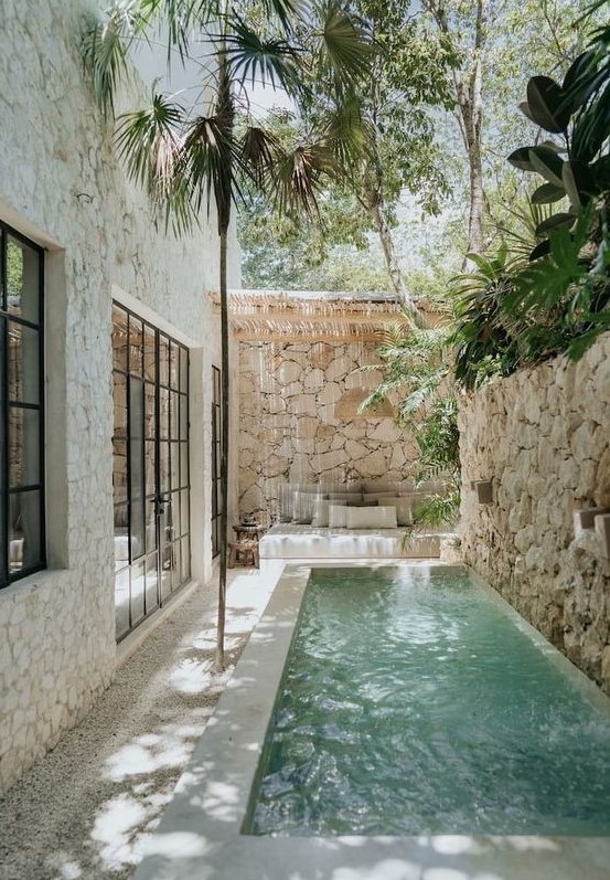 a tropical outdoor space with a stone fence, a narrow pool with a waterfall, a seat with pillows and some trees over the space
