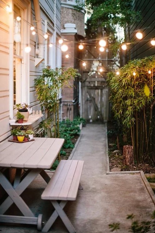 a side yard with a concrete path, a wooden dining set, bamboo and greenery and string lights