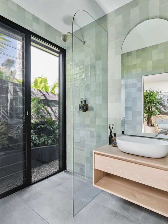 a small modern bathroom with a glass door that leads to the garden, green zellige tiles, a floating vanity and an arched mirror