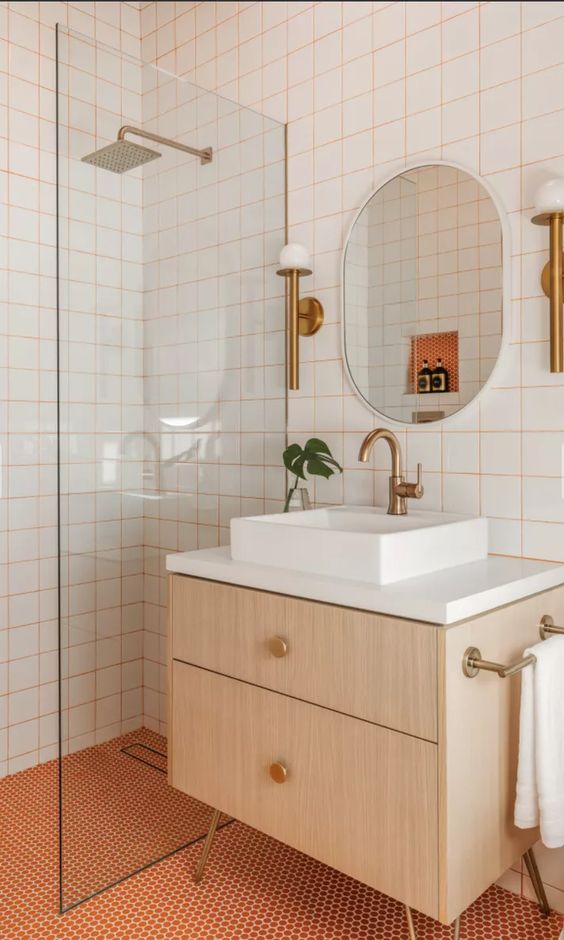 a bright modern bathroom with white square tiles with orange grout and orange penny tiles, a timber vanity, an oval mirror and brass fixtures