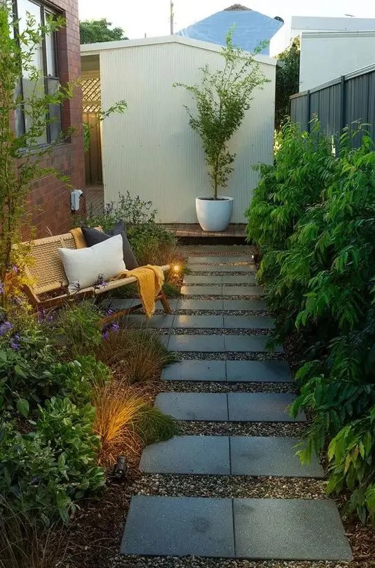 a long and narrow side yard with a tile pathway, greenery, grasses and blooms, a rattan bench with pillows and some lights