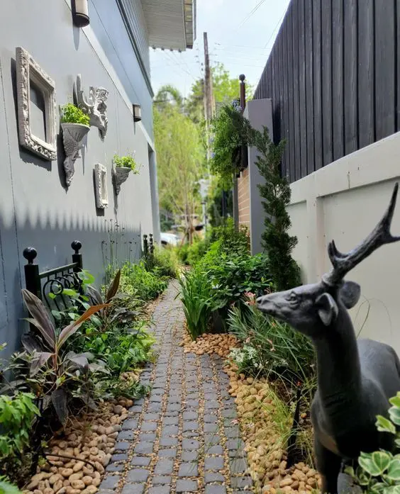 a lovely and creative side yard with a stone path and pebbles, greenery and shrubs, a black forged bench, a black deer and frames and plants on the wall