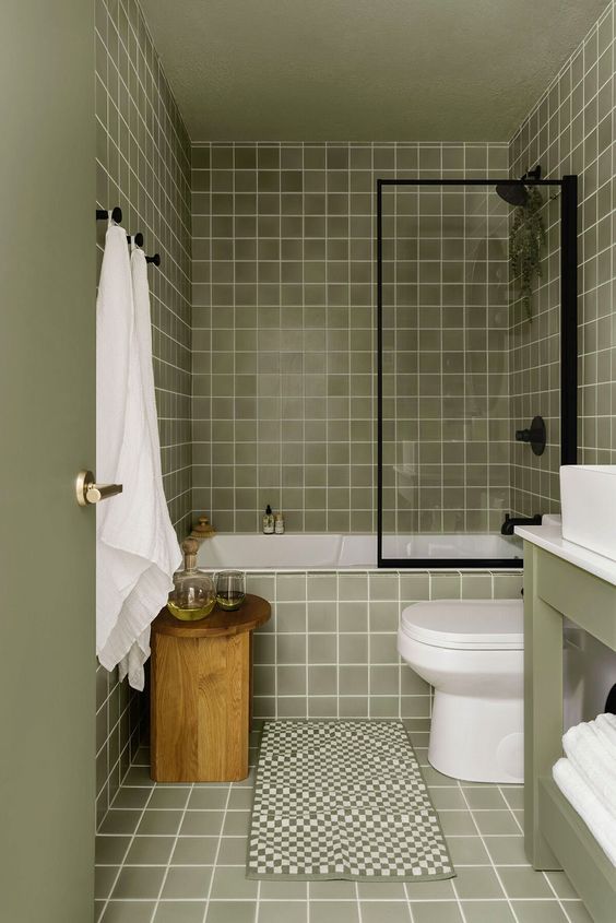 a light green bathroom clad with square tiles, a green vanity, a wooden stool, black fixtures and white appliances