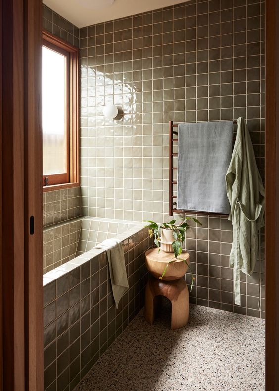 a modern bathroom with a terrazzo floor, green square tiles and a tub clad with tiles, timber and stained wood plus a wooden stool