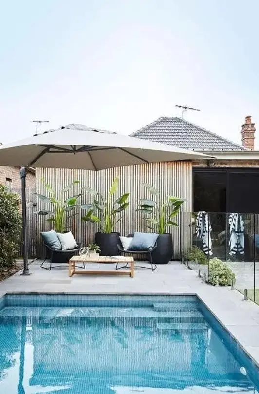 a pretty tropical backyard with a blue pool, a parasol, potted plants and a coffee table, greenery and a stone deck
