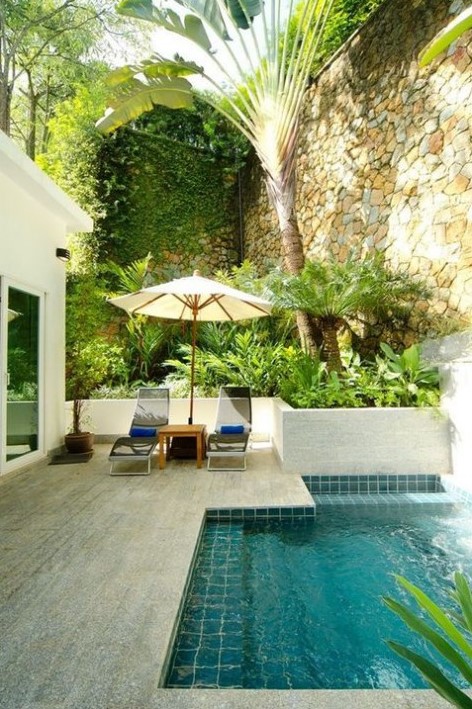a stone clad pool deck with loungers and an umbrella plus a small backyard pool clad with turquoise tiles inside