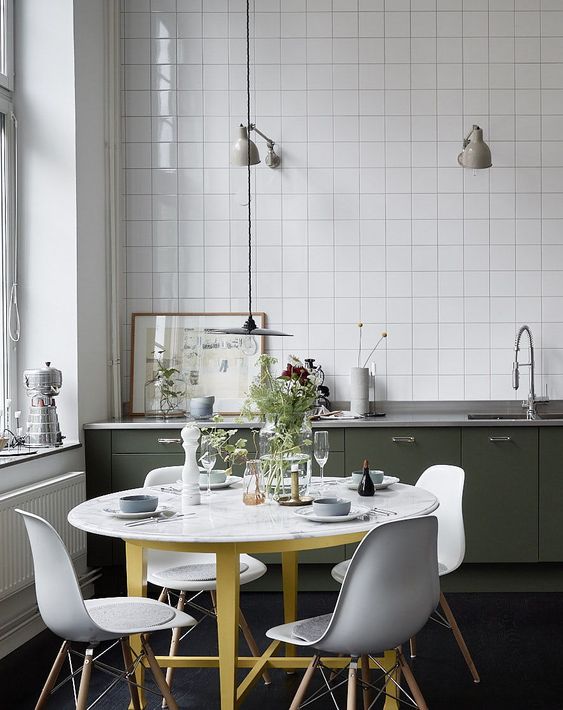 a pretty Nordic kitchen with dark green lower cabinets, white square tiles, a round table and white Chairs and grey lamps