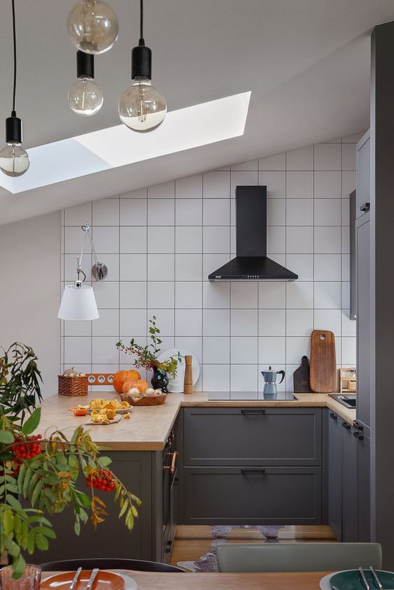 a small attic kitchen with graphite grey lower cabinets, a white square tile backsplash, a black hood and greenery