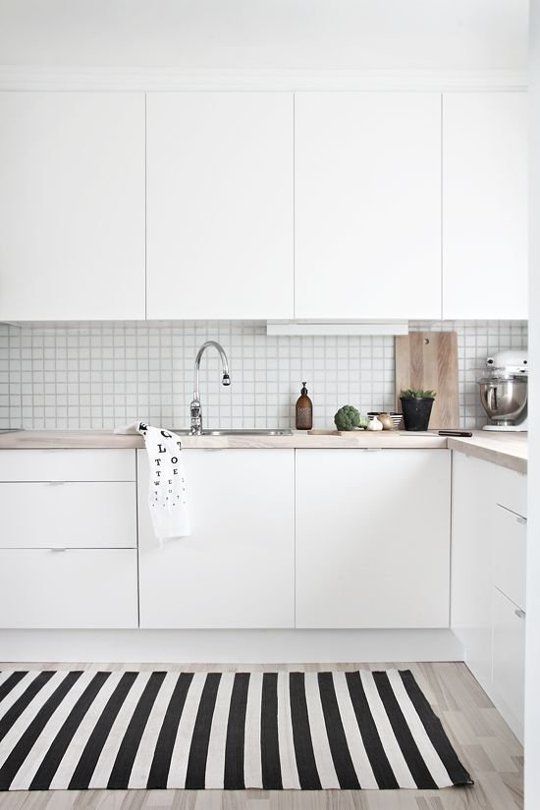a white Scandinavian kitchen with sleek cabinetry, a square tile backsplash and butcherblock countertops, a striped rug