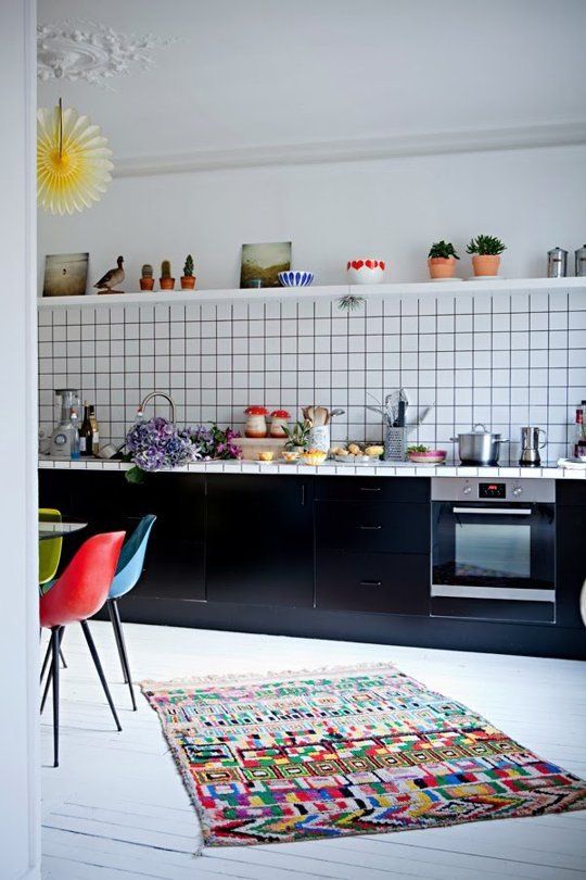 a bold kitchen with black cabinets and a white square tile backsplash, a bold rug and colorful chairs at the table