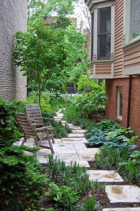a welcoming side yard with pavements, lots of various greenery and shrubs and a tree, some wooden chairs