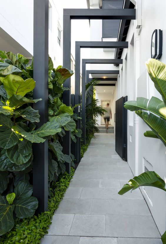 An ultra modern side yard with a tiled path, greenery and tropical plants and some black wooden beams over the path