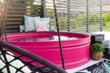 a bold patio with a hot pink stock tank pool, a black ladder, a woven chair with a pillow and some more pillows on the bench