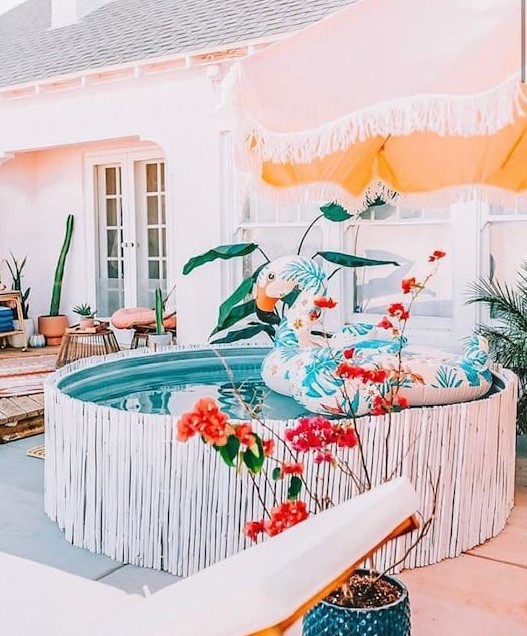 a bright and cool backyard with a wooden deck, a stock tank pool covered with whitewashed sticks, a colorful float, an umbrella and bold potted blooms