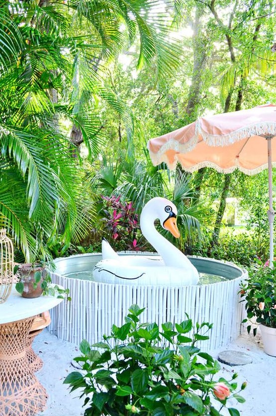 a bright and funny pool nook with a stock tank pool clad with a stick cover, potted greenery all around, a pink umbrella and rattan furniture