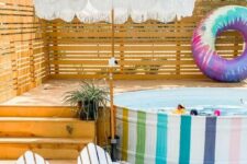 a bright outdoor nook with a rainbow stock tank pool, a wooden deck, colorful floats, an umbrella, white chairs and a wooden rug