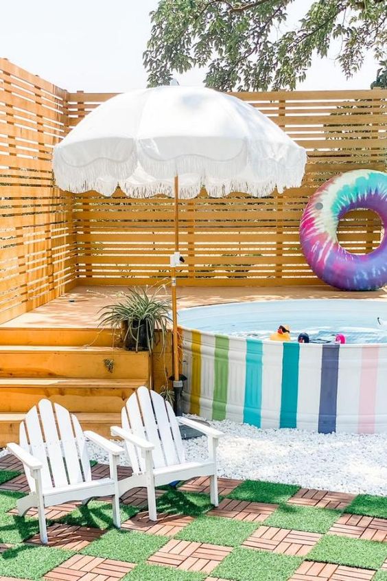 a bright outdoor nook with a rainbow stock tank pool, a wooden deck, colorful floats, an umbrella, white chairs and a wooden rug