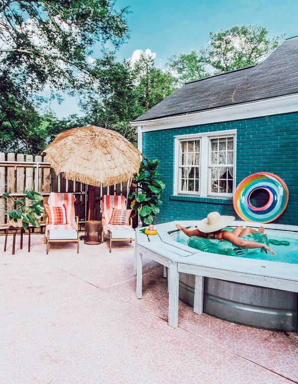 a cool and bright outdoor space with pink large format tiles, a stock tank pool with a wooden deck around it, loungers, an umbrella and bright floats