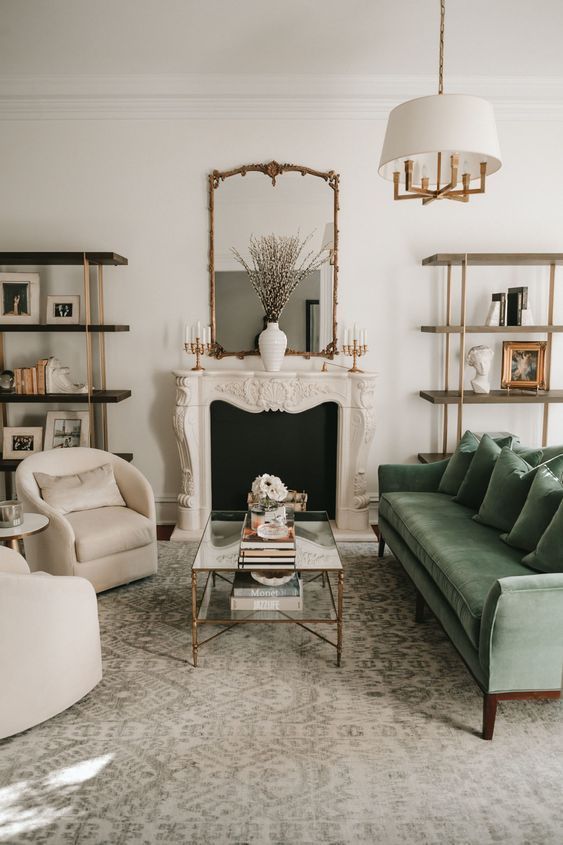 a cozy neutral living room with a faux fireplace, bookshelves, neutral chairs, a green sofa, a glass coffee table, a printed rug and a pendant lamp