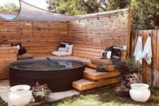 a cozy outdoor space with a stepped wooden deck with pillows, a black stock tank pool, a cover and lights