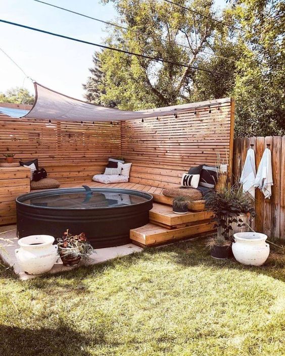 a cozy outdoor space with a stepped wooden deck with pillows, a black stock tank pool, a cover and lights
