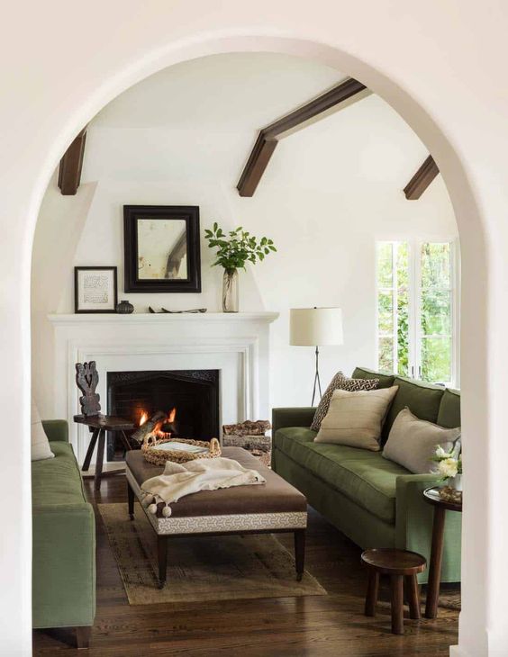 a lovely Mediterranean living room with two green sofas, a fireplace, an ottoman, some side tables, wooden beams on the ceiling