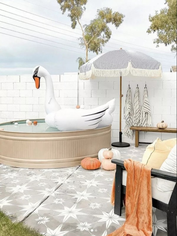 a lovely outdoor space with a tiled deck, a stock tank pool, wooden chairs and benches, an umbrella and some towels