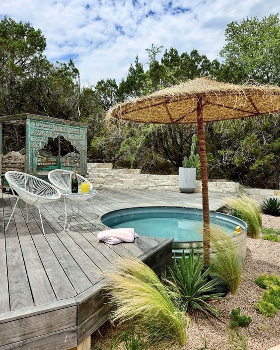 a lovely outdoor space with a wooden deck, a stock tank pool, white chairs, an umbrella and some greenery