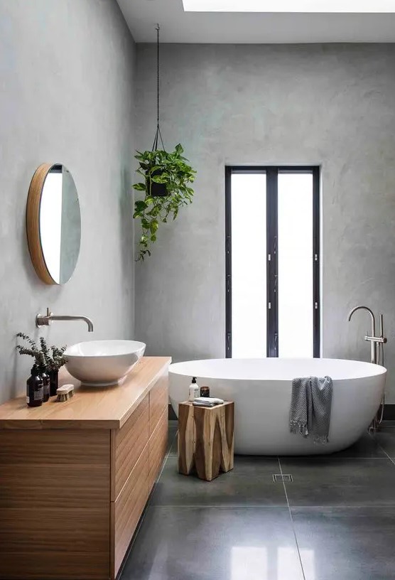 a minimalist grey bathroom with concrete walls and large scale tiles on the floor, a stained vanity, an oval tub, potted greenery and a window
