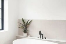 a minimalist serene bathroom clad with greige tiles, with an oval tub, black fixtures and a potted plant plus a window to enjoy natural light