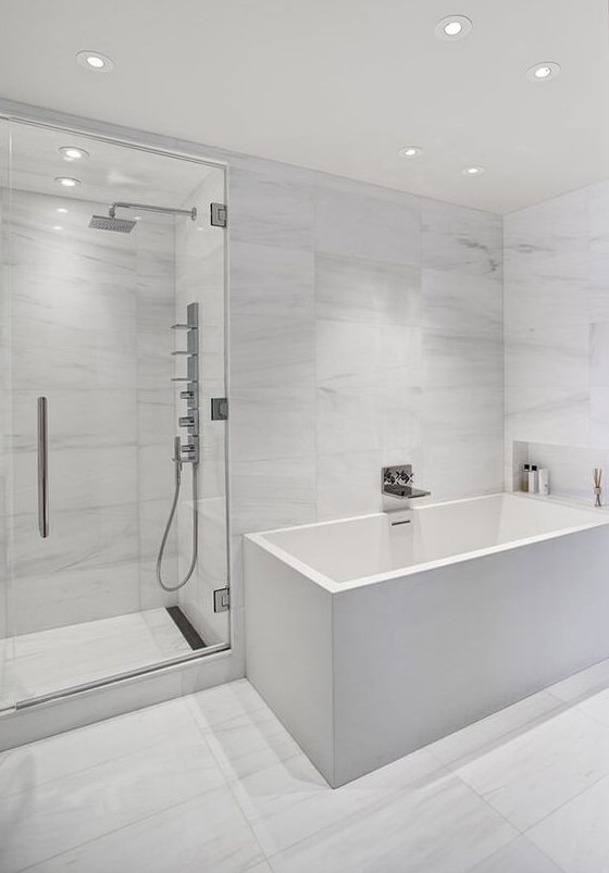 A minimalist white bathroom with marble large scale tiles, a square tub and built in lights all over
