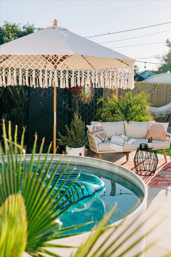 a modern boho space with a tiled deck, a stock tank pool, an umbrella with tassels, a sofa with pillows