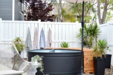 a modern stock tank pool in black, a wooden deck, an umbrella, potted greenery, a woven chair and a side table