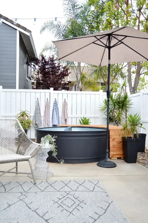 a modern stock tank pool in black, a wooden deck, an umbrella, potted greenery, a woven chair and a side table