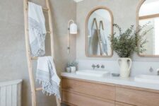 a neutral bathroom with a wooden floating vanity, arched mirrors in wooden frames, a ladder and neutral textiles