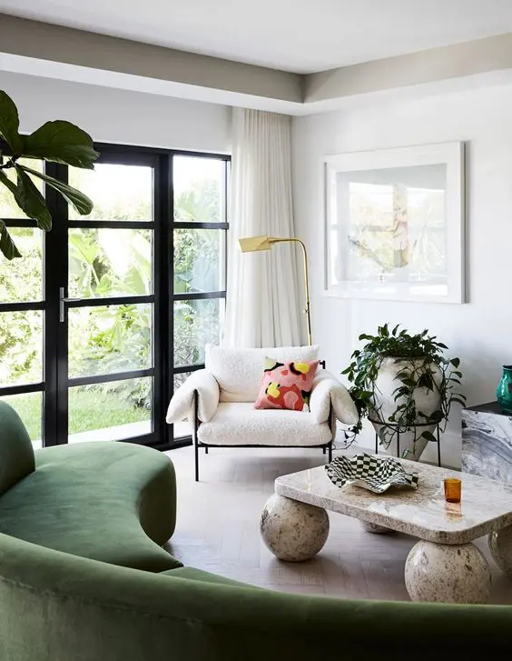a neutral living room with a curved green sofa, a neutral chair, a stone coffee table and side table and plants