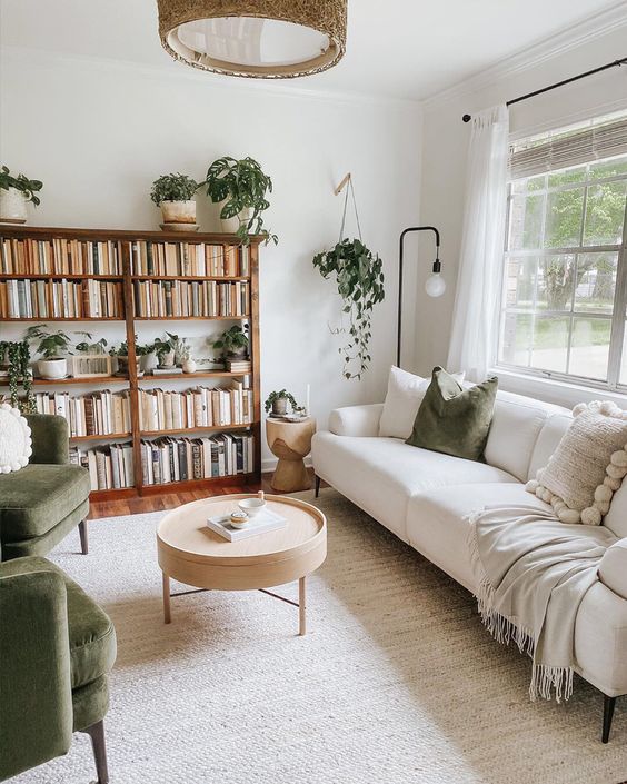 a neutral living room with a stained bookshelf, a creamy sofa, green chairs and pillows and lots of potted plants