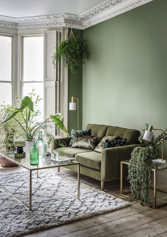a refined living room with an olive green wall, a moody green sofa, some glass tables and lots of greenery