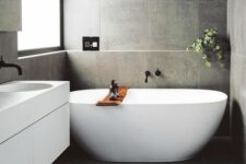a refined minimalist bathroom with grey large scale tiles, an oval tub and a white vanity, a window and some greenery