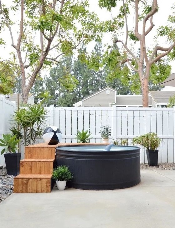a simple and pretty backyard with a black stock tank pool, a wooden ladder and a deck, potted plants and fun floats is welcoming