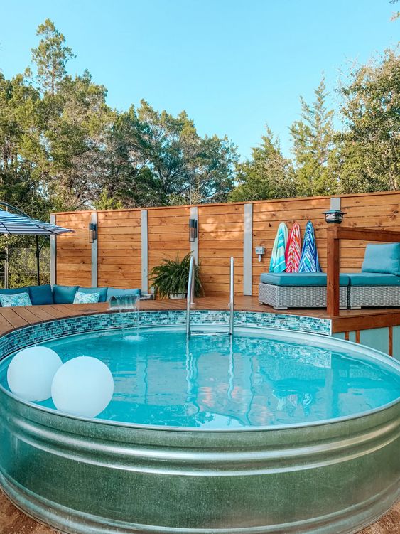 a stock tank pool with floats, a wooden deck with a daybed, some greenery and bright towels